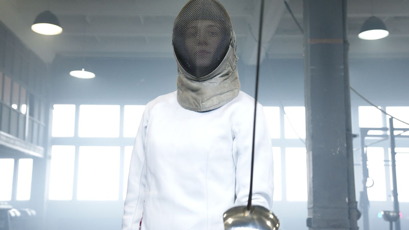 A fencer standing en garde with épée upright in a hall with tall windows.